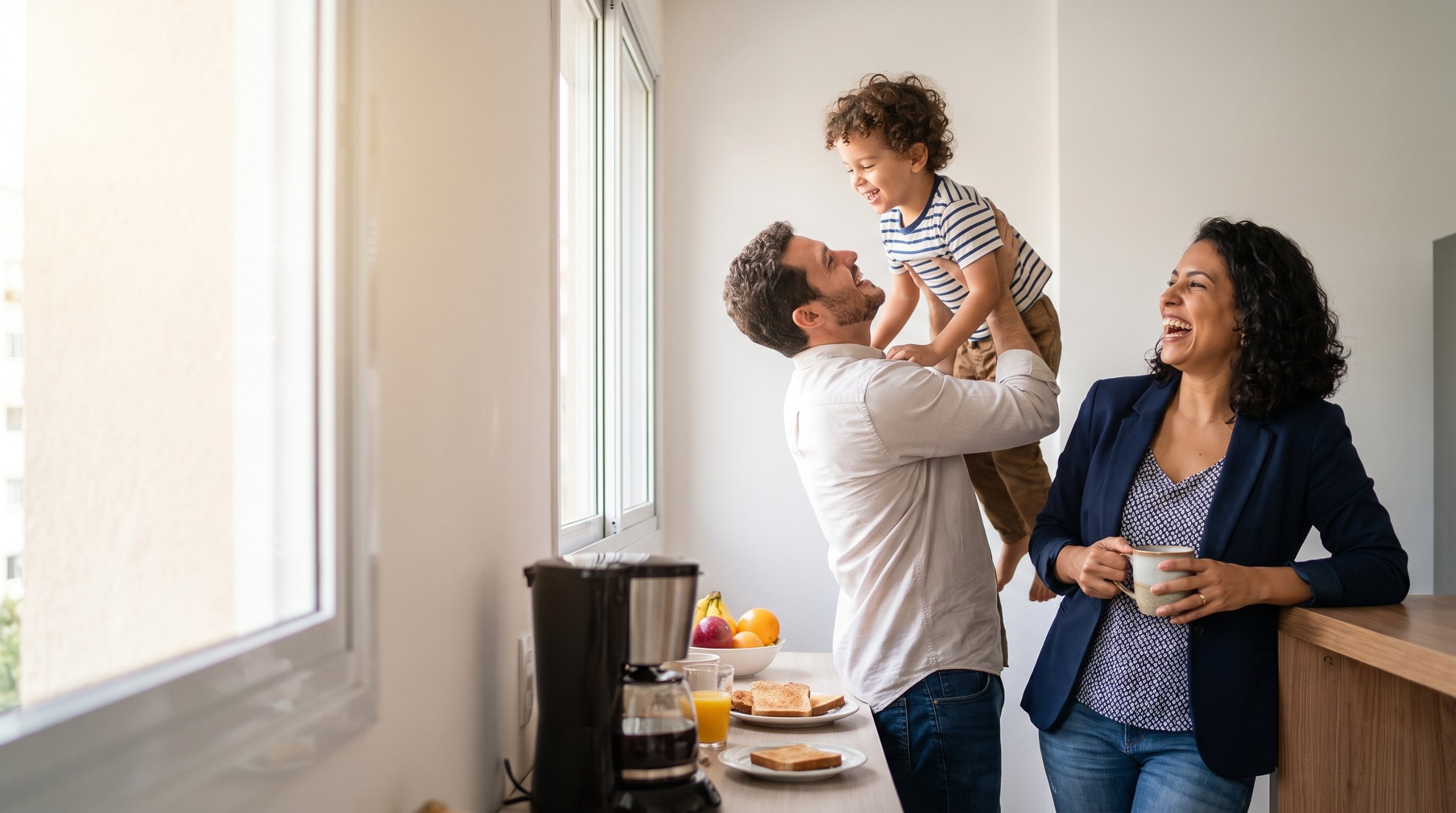 Família na cozinha de casa pela manhã, o pai levantando o filho no colo.